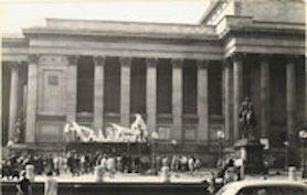 Large inflatable sculpture in front of civic building with a crowd of people and street traffic